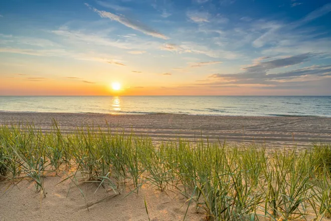 Die Sonne am Meereshorizont, von einem Strand aus gesehen, mit Dünengras im Vordergrund.